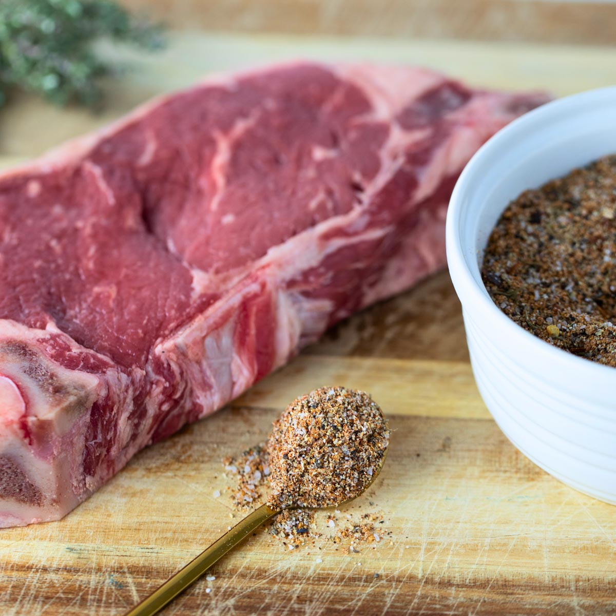 spoon of steak seasoning sitting on wooden cutting board next to steak and bowl of steak seasoning