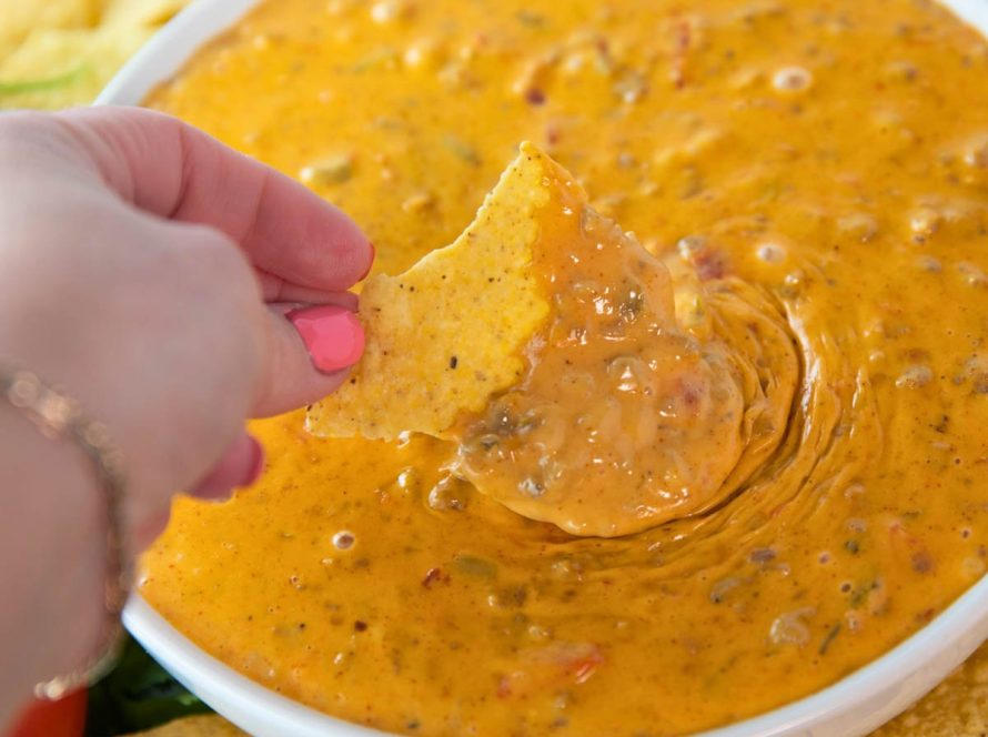 woman's hand holding chip and scooping easy rotel dip with sausage. Bowl is surrounded by tortilla chips.